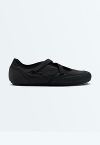 Chaussure plate style Mary Jane noire avec tissu matelassé, bout arrondi et bride Velcro sur le dessus du pied sur fond uni.