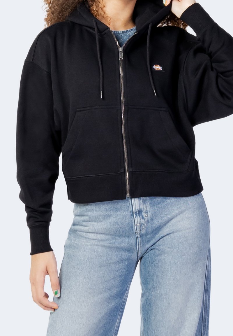 Personne portant un sweat à capuche noir zippé avec poches devant et un jean bleu, montrée des épaules aux hanches, main touchant la capuche.
