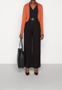 Svarta vida byxor med veck, parat med en svart topp med djup V-ringning och en orange cardigan. En svart handväska fulländar outfiten.
