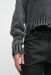 Pulôver cinza de malha com uma bainha desfiada e mangas texturizadas, apresentando grandes botões pretos. Usado sobre jeans pretos rasgados.