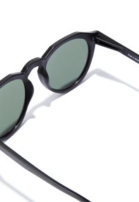 Ochelari de soare negri cu lentile rotunde, nuanță verde și rame unghiulare. Au brate negre drepte cu un branding subtil pe o parte.