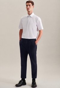 Camicia bianca a maniche corte con bottoni e taschino sul petto, abbinata a pantaloni sartoriali blu navy, indossata con scarpe nere e calzini.