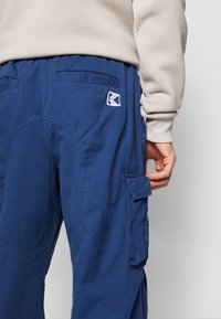 Mørkeblå cargo-bukser med lommedetaljer, elastisk midjebånd og et lite logo-patch. Stoffet virker slitesterkt med en jevn tekstur.