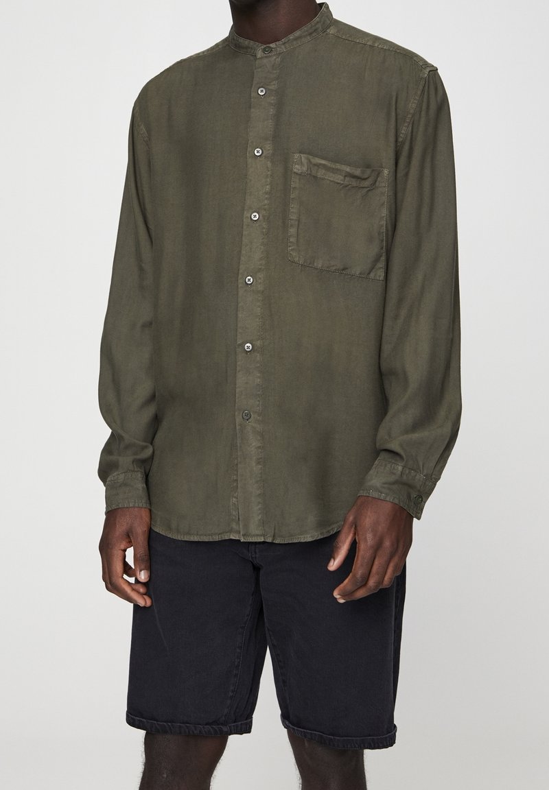 Homme portant une chemise olive à manches longues avec poche poitrine et un short noir à ourlet roulé, debout devant un fond uni.