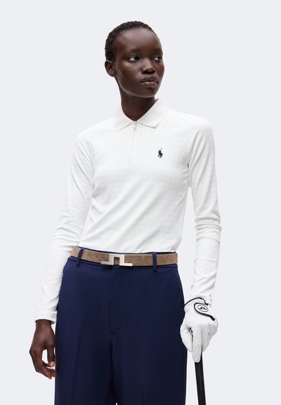 Femme portant un polo blanc à manches longues, un pantalon bleu marine avec une ceinture beige et un gant de golf blanc tenant un club de golf.