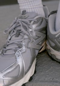 Zapatillas deportivas grises con parte superior de malla, cordones y suelas de goma. Presentan un logotipo prominente y acentos texturizados en los costados.