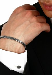 Silverfärgad kedjebransolet med en slät rektangulär lås, bärs på en hand med en synlig vit manschett. Texturerade metallaccenter på manschetten.
