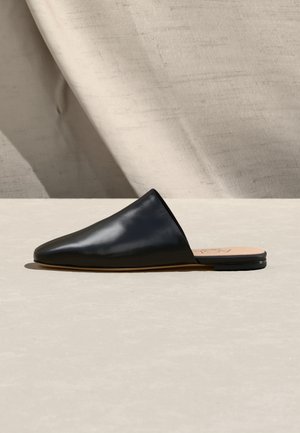 Black pointed-toe leather slip-on flat with open back on a light beige surface and fabric backdrop.