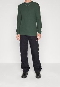 Grüner strukturierter Strickpullover mit Rundhalsausschnitt, kombiniert mit schwarzen Cargo-Hosen mit Seitentaschen und schwarzen Schnürschuhen.