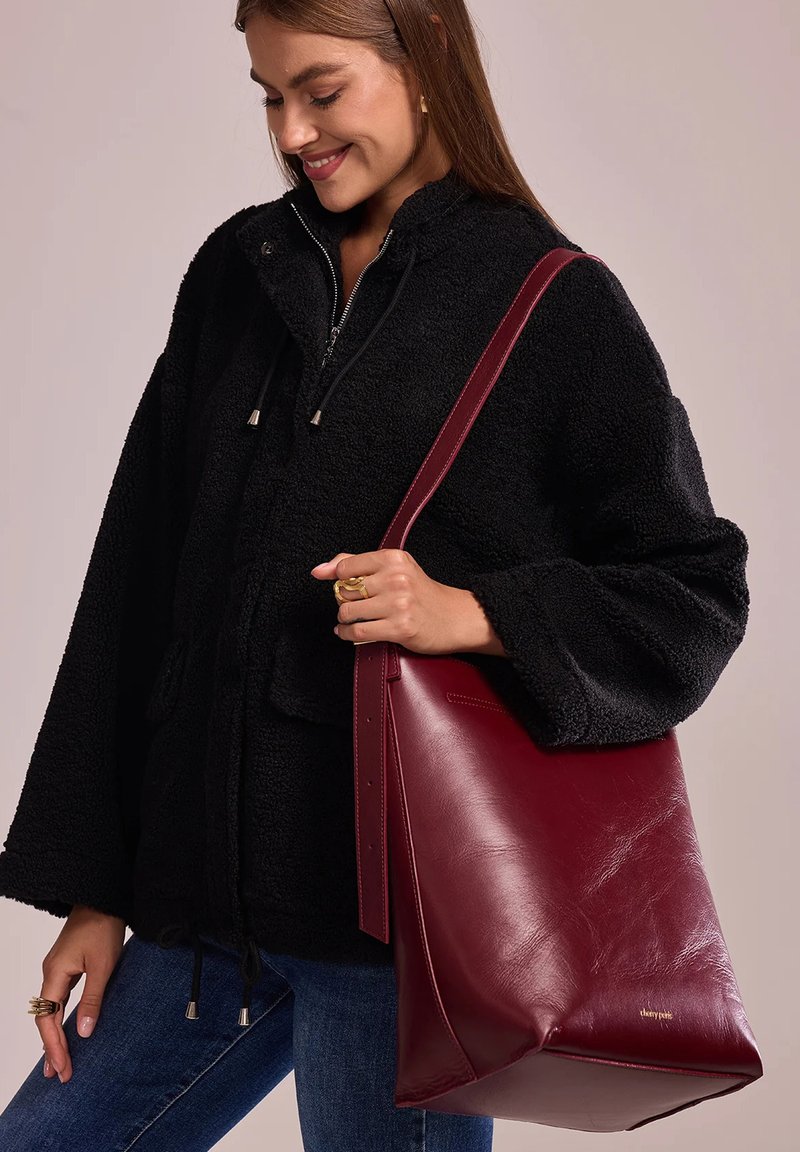 Sac fourre-tout en cuir bordeaux au design structuré, texture lisse et sangle réglable. Associé à une veste en polaire noire et à un jeans.