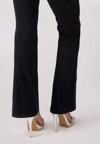Czarne spodnie z rozszerzanym dołem, w połączeniu z białymi sandałami na wysokim obcasie, które mają beżową wkładkę i srebrne akcenty na obcasach. Gładki materiał, smukła sylwetka.