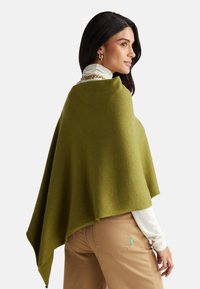 Poncho lavorato a maglia verde con una vestibilità comoda, caratterizzato da una forma triangolare, una texture morbida e bordi arrotolati, indossato su una maglietta bianca e pantaloni beige.