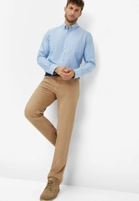Hellblaues Hemd mit Knöpfen und Brusttasche, kombiniert mit khakifarbenen Chinos und braunen Schuhen, vor einem schlichten weißen Hintergrund.