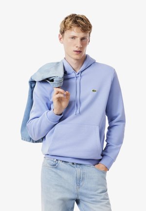 Sudadera de color púrpura claro con bolsillo canguro, cordones y un pequeño logo verde. El modelo lleva una chaqueta de mezclilla sobre un hombro y viste jeans claros.