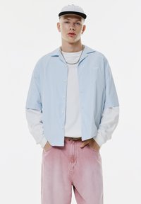 Jeune homme à la peau claire portant une casquette blanche, une chemise à rayures blanches et bleu clair superposées, un pantalon rose et un collier en chaîne argenté, les mains dans les poches.