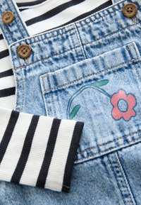 Denim-overalls har en børstet finish med træknapper, suppleret med en langærmet, stribet bomuldstrøje og blomsterbroderi på lommen.