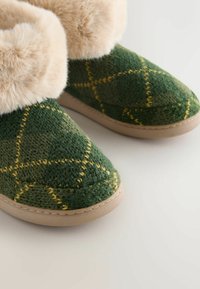 Papuci tricotati verzi cu o margine pufoasă de culoare bej, având un model în diamant de culoare galbenă și o talpă deschisă. Textură moale, formă rotundă.