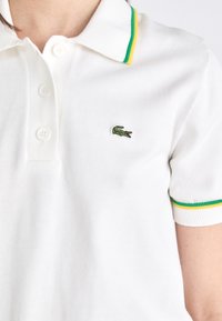Witte polo met drie knopen, groen en gele gestreepte kraag en mouwboorden, met een klein groen krokodillenlogo.