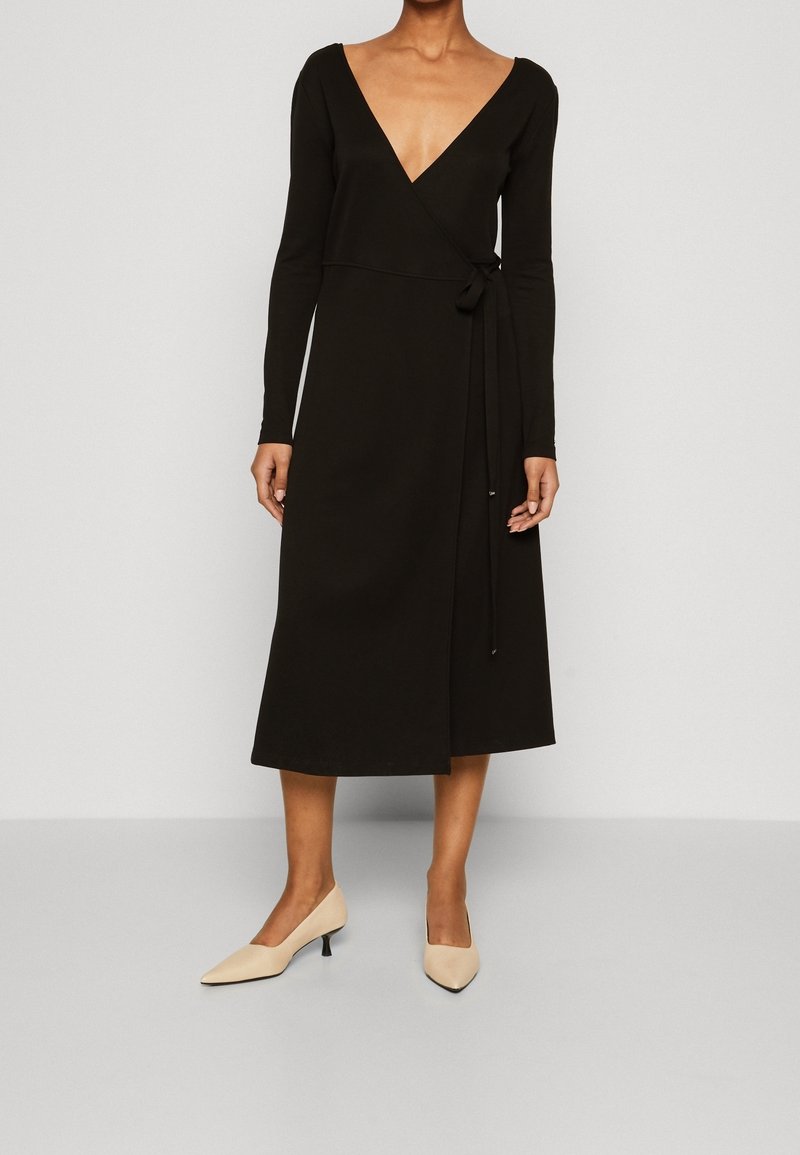 Femme portant une longue robe portefeuille noire à manches longues et col en V, associée à des chaussures pointues beiges à talons bas sur un fond uni.