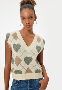 Strikket ærmeløs sweater med V-hals, der har en cremefarvet bund, grønne og brune hjerte- og diamantmønstre samt ribbet kant.