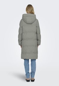 Long manteau gris matelassé avec une capuche spacieuse, présentant un design matelassé et des poignets élastiques, porté sur un jean bleu et des chaussures grises.
