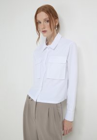Donna con capelli rossi che indossa una camicia bianca con bottoni, dotata di due tasche frontali, e pantaloni beige a vita alta plissettati.