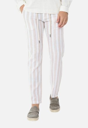 Persona che indossa pantaloni beige e bianchi a righe verticali con coulisse e scarpe slip-on grigie, in piedi davanti a uno sfondo bianco.