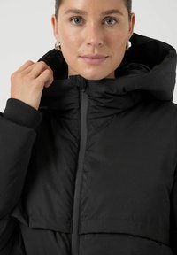 Giacca imbottita nera con collo alto, chiusura con cerniera e tessuto strutturato. Presenta polsini a coste e un cappuccio per una maggiore protezione dal freddo.