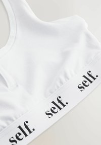 Valkoinen puuvillainen bralette, jossa on sileä pinta, ja leveä elastinen nauha, johon on painettu "self." mustalla. Yksinkertainen muotoilu ja urheilurintaliivimäinen selkä.