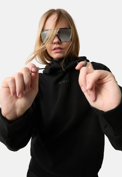 Schwarzer Pullover mit hohem Kragen, auf dem der Markenname "FIRE + ICE" in erhabenem Text gestaltet ist. Das Modell trägt große, reflektierende Sonnenbrillen.