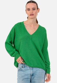 Femme aux cheveux tirés en arrière, portant un pull vert à col en V ample et un jean bleu clair, debout sur un fond blanc uni.