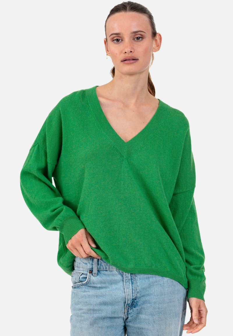 Femme aux cheveux tirés en arrière, portant un pull vert à col en V ample et un jean bleu clair, debout sur un fond blanc uni.