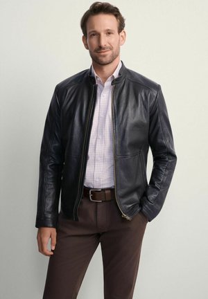 Mężczyzna w czarnej skórzanej kurtce na jasnej, kraciastej koszuli i brązowych spodniach, stojący z jedną ręką w kieszeni na jednolitym tle.