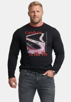 Blondyn w czarnej koszulce z długim rękawem, z grafiką krętej drogi i czerwonym tekstem, który mówi "Catch us In the Pits."