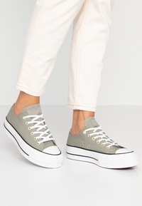 Light green canvas sneakers with white rubber soles and toe cap. Features white laces and gold eyelets; black accent stripe along the sole.