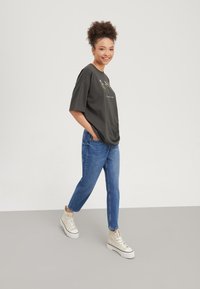 Giovane donna che cammina con le mani nelle tasche, indossando una t-shirt nera oversize, jeans blu e sneaker alte crema su pavimento grigio.
