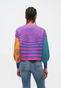 Persona ar pītām matu bizēm, valkājot daudzkrāsainu džemperi ar rozā, zilu, oranžu un zaļu rakstu motīviem un gaiši zilas džinsas, redzama no muguras.