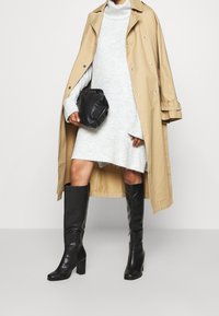 Trench beige sopra un vestito a maglia grigio chiaro, abbinato a stivali neri con tacco alti fino al ginocchio e una borsa nera con texture.