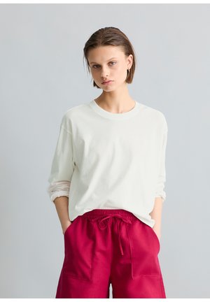 Jonge vrouw met kort haar, gekleed in een wit shirt met lange mouwen en een high-waist rode broek met trekkoord, staand met haar handen in de zakken.