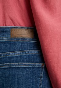 Jean en denim bleu avec une étiquette en cuir marron "ESPRIT Denim", porté avec un haut rose texturé partiellement rentré.