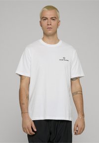Camiseta de algodón blanca con mangas cortas y cuello redondo. Presenta un pequeño logo negro en el pecho. Diseño sencillo y de ajuste relajado.