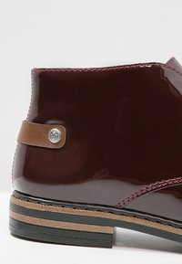 Bottine en cuir bordeaux foncé brillant avec languette en cuir marron et semelle superposée noire et beige, coutures visibles le long des bords.