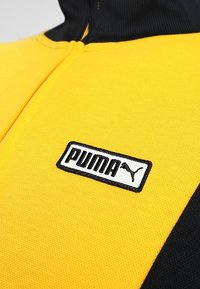 Puma träningsjacka med en gul och svart design, texturerad tyg, halvmåttslång dragkedja och en sytt logotyp patch på bröstet.