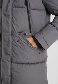 Cappotto isolante grigio con superficie testurizzata, cuciture orizzontali e tasca frontale con chiusura a scatto e dettaglio con zip.