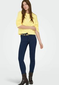 Pull en tricot jaune, coupe courte, associé à un jean skinny bleu foncé et des bottines noires à fini texturé.