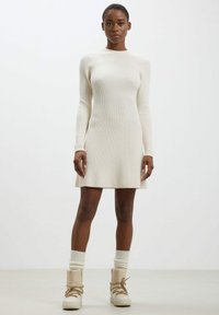 Abito lavorato a maglia di colore crema con maniche lunghe, texture a coste e vestibilità ampia. Abbinato a stivali beige con lacci e calze lavorate a maglia di colore crema.