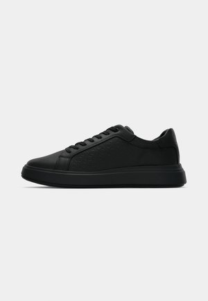 Schwarze Low-Top Sneakers mit tonalem "CK"-Muster, schnürbarem Design und dicker Sohle auf einem weißen Hintergrund.