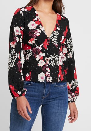 Kvinde iført en sort langærmet bluse med røde og hvide blomster, kombineret med blå jeans, stående mod en ensfarvet baggrund.
