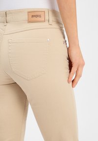 Beige rekbare spijkerbroek met een platte achterzak en een tan leren logo-applicatie met het label "ANGELS" op de tailleband. Gladde textuur en een aansluitend ontwerp.