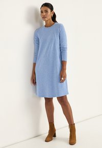 Hellblaues Pulloverkleid mit langen Ärmeln, gerippter Struktur, geschwungenem Saum und lockerem Sitz, kombiniert mit beigefarbenen Stiefeletten mit Absatz.